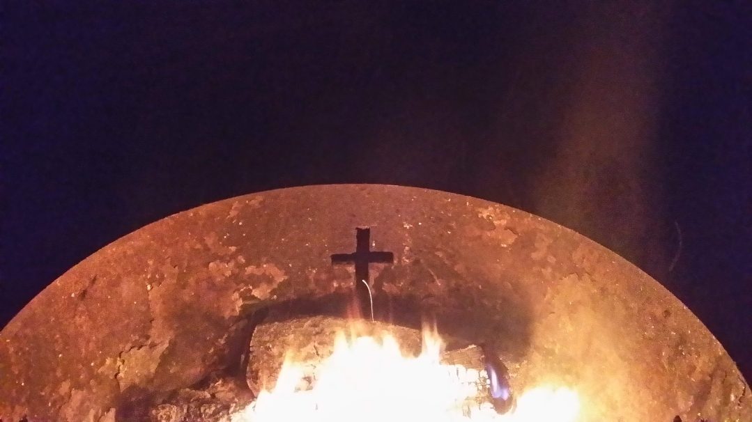 The fire ring at night, a cross cut into the steel glowing above the flames