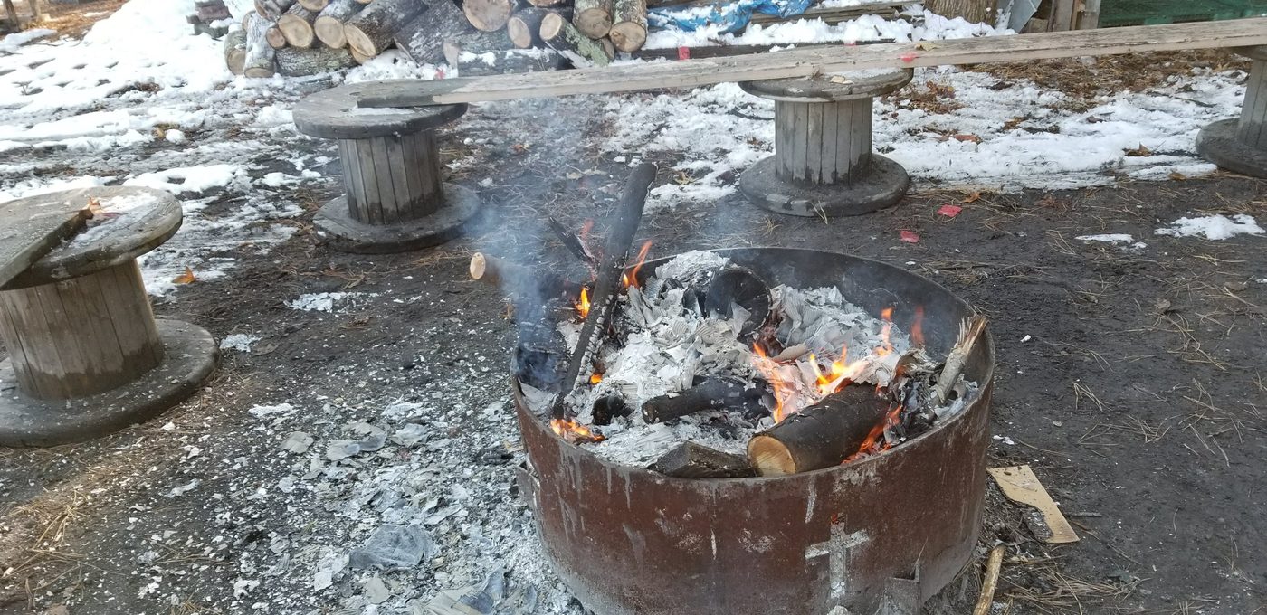 The fire ring burning in the snow, a cross cut into the steel, with cable-spool benches around it
